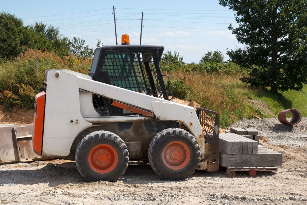 A White and Orange Skid Steer is Parked in a Dirt Field — Grafton Concreting & Bobcat Services In South Grafton, NSW