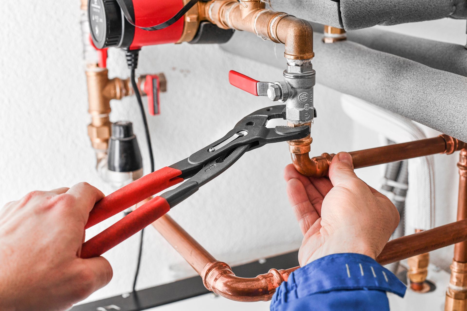 Hands using red-handled pliers to tighten a copper pipe under a sink