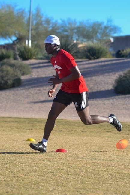 Runner in red shirt sprinting on a grassy field near orange training cones.