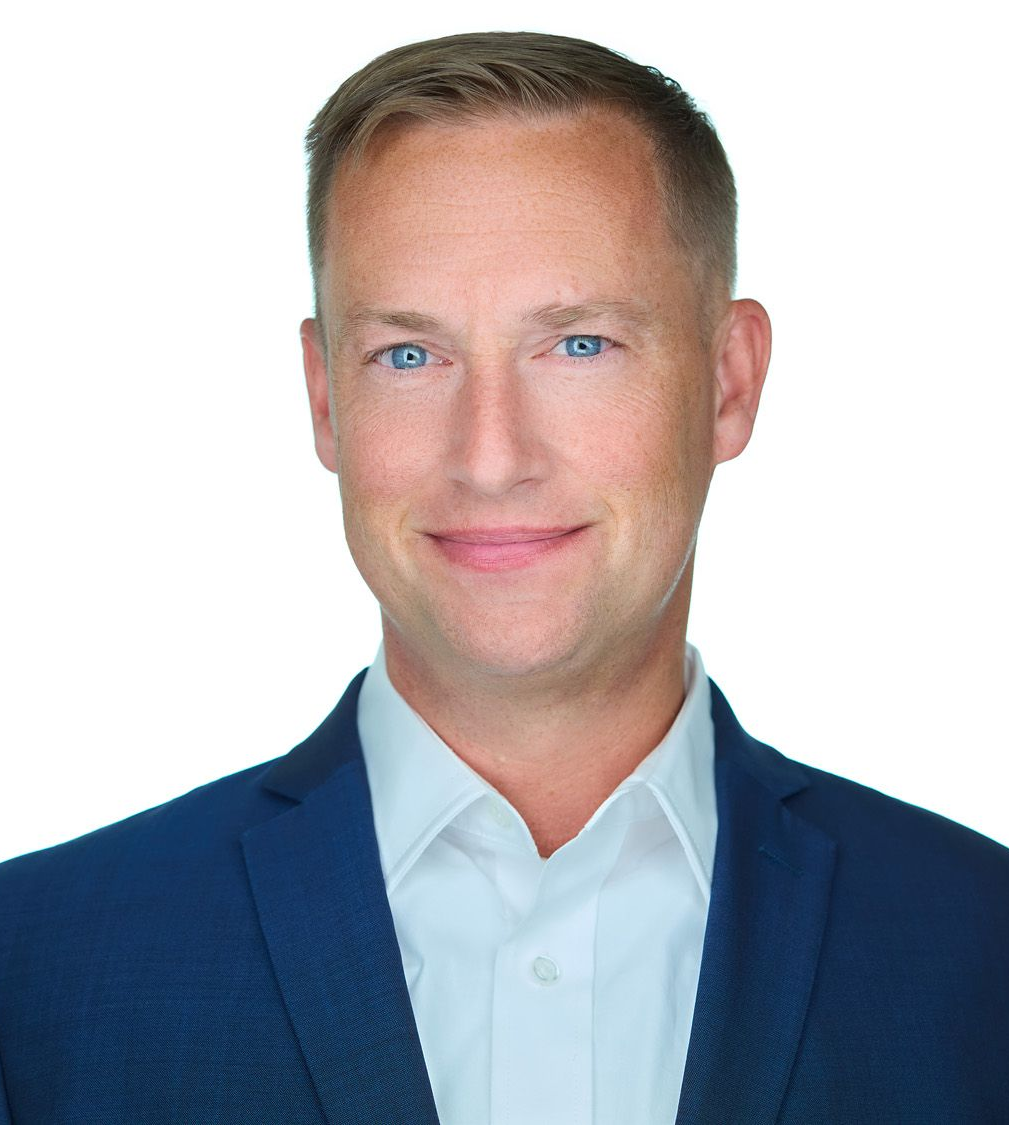 Man in a blue blazer and light blue shirt smiles, headshot against a white background.