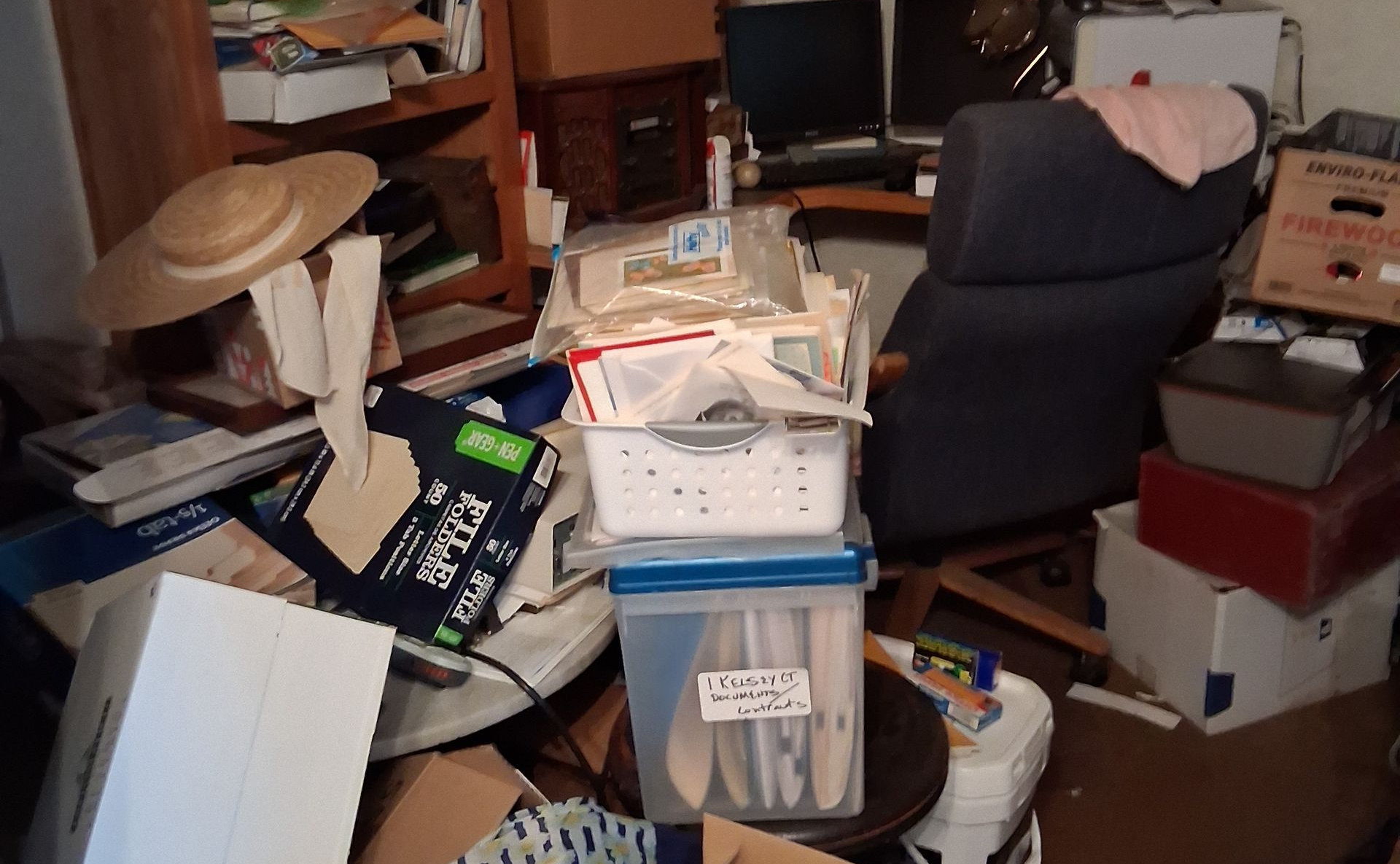 Cluttered home office with stacked boxes, papers, a chair, and hats on furniture in a messy room