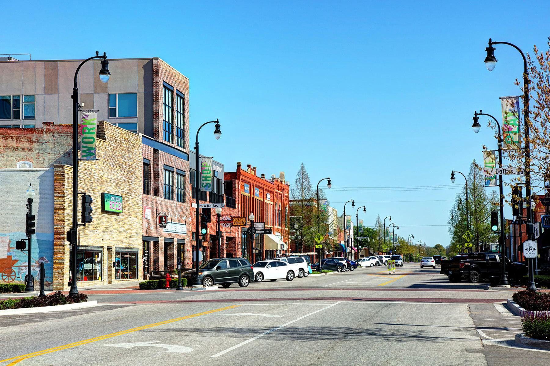 Sunny small-town street with brick buildings, parked cars, and lampposts under a clear blue sky