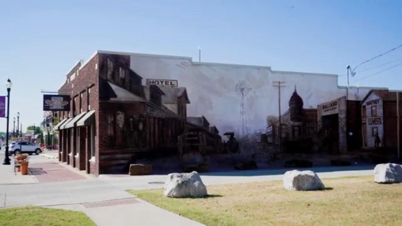 Street view of a weathered brick building with mural, storefronts, and a grassy foreground with rocks