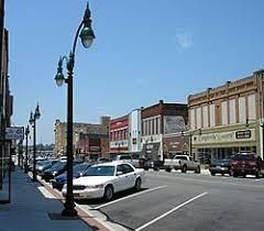 Small-town main street with parked cars, storefronts, and vintage streetlamps under a clear blue sky