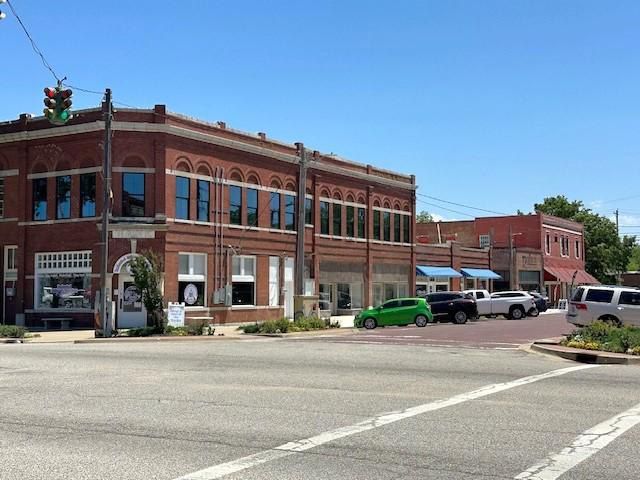 Brick corner storefronts on a sunny street intersection, with parked cars and a traffic light.