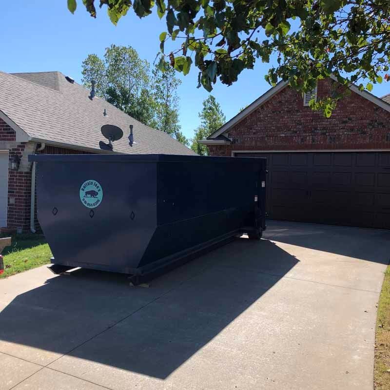 Large black dumpster parked in a suburban driveway beside a brick garage on a sunny day