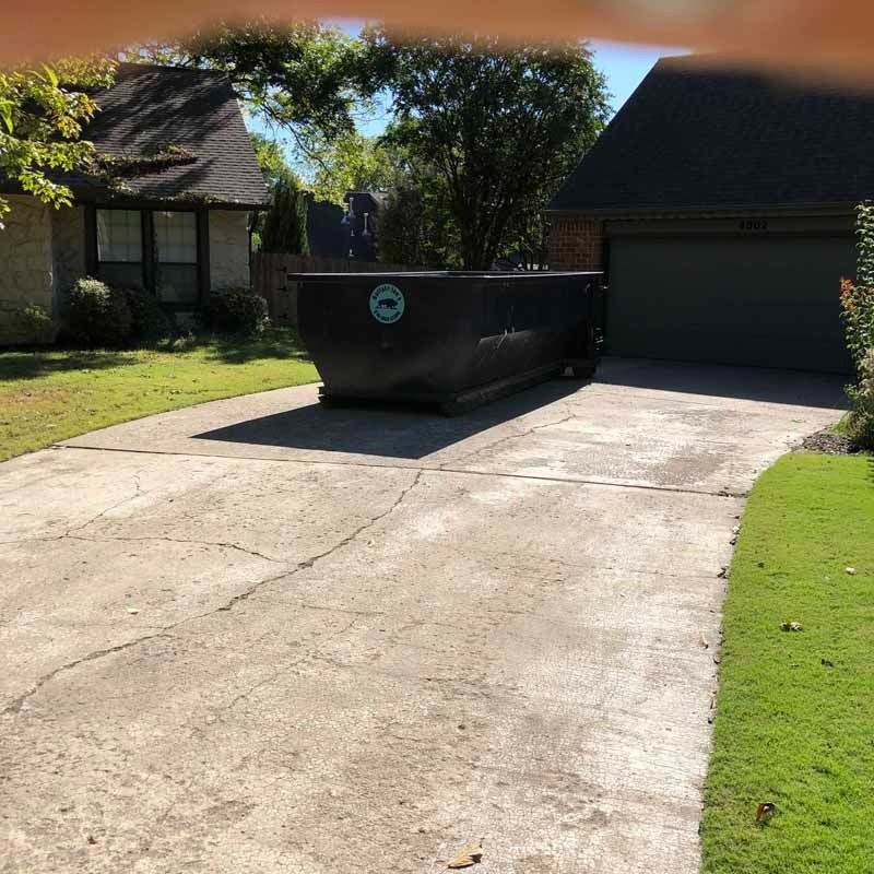 Black dumpster on a driveway beside a garage in a sunny suburban yard