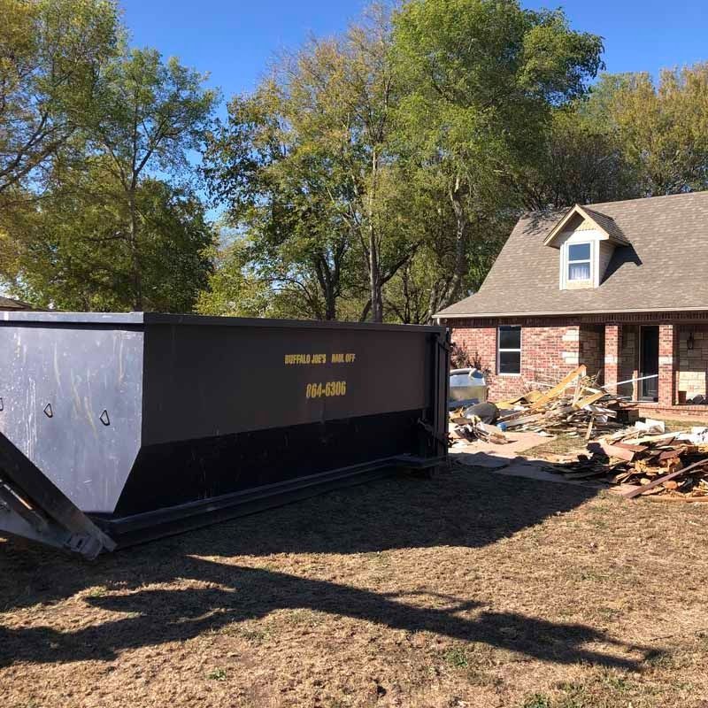 Large dumpster beside a house amid demolition debris in a yard under sunny blue skies