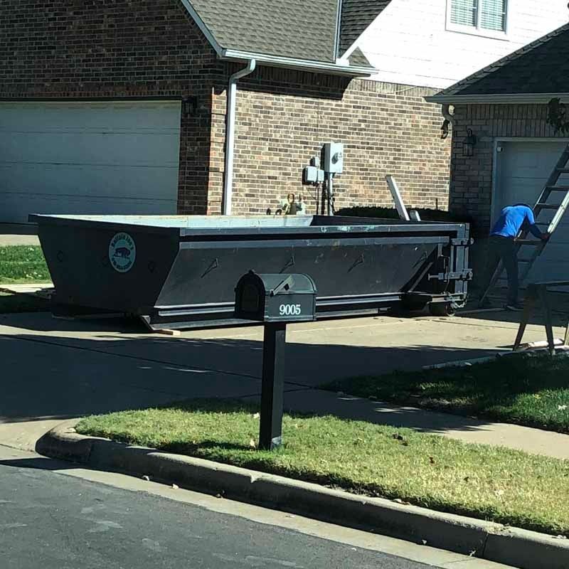 Black dumpster trailer parked in a suburban driveway beside a brick house and mailbox
