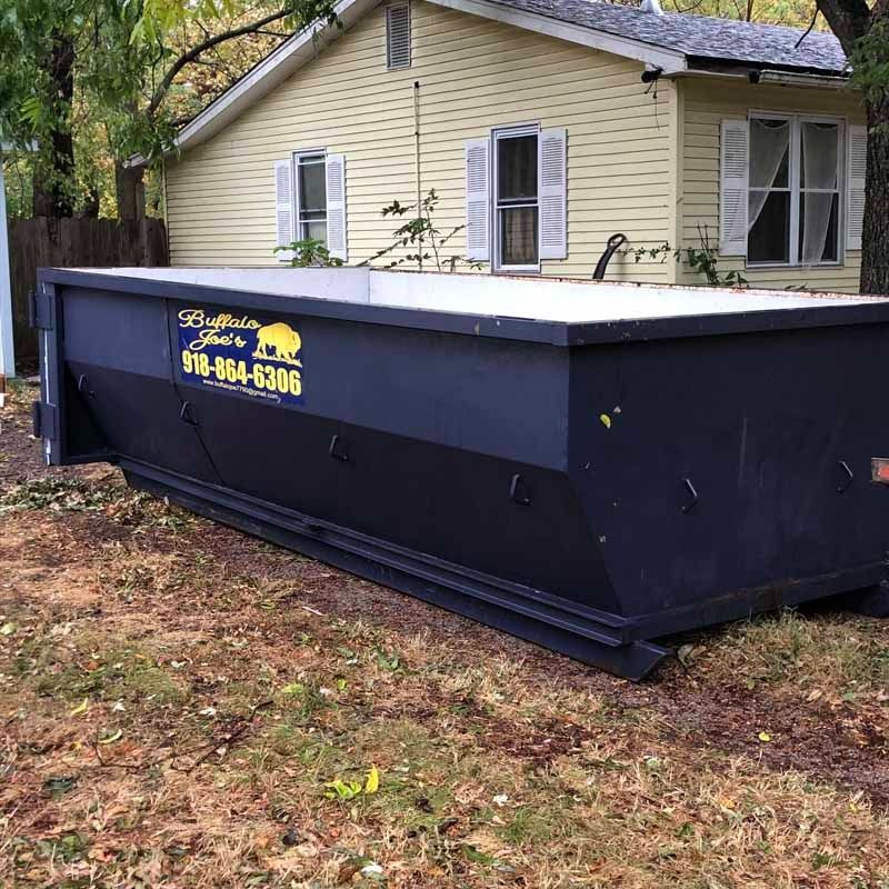 Large black dumpster beside a beige house in a wooded yard