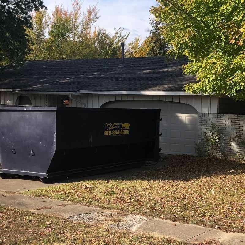 Black dumpster in front of a gray house with a dark roof and autumn trees behind it