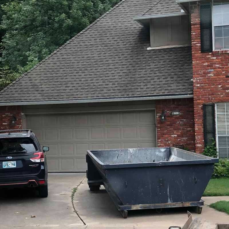 Two cars in a driveway beside a garage, with a large black dumpster in front.
