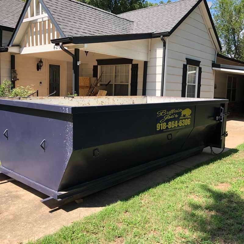 Blue dumpster on a driveway in front of a white house with a porch and dark roof.