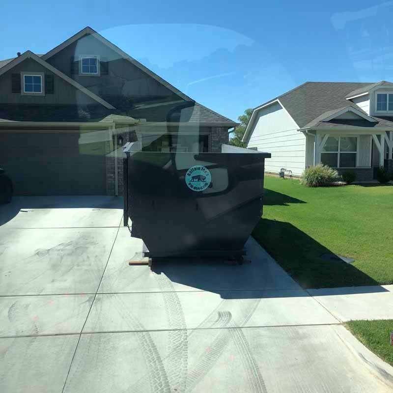 Suburban driveway with a large black dumpster in front of a house, viewed through a windshield.