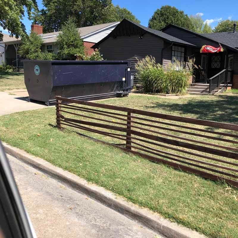 Residential house with a large dumpster in the driveway and a brown metal fence along the curb