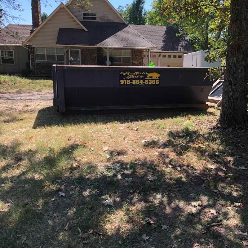 Dark dumpster in front of a house, on a grassy lawn. The dumpster has a yellow logo and phone number.