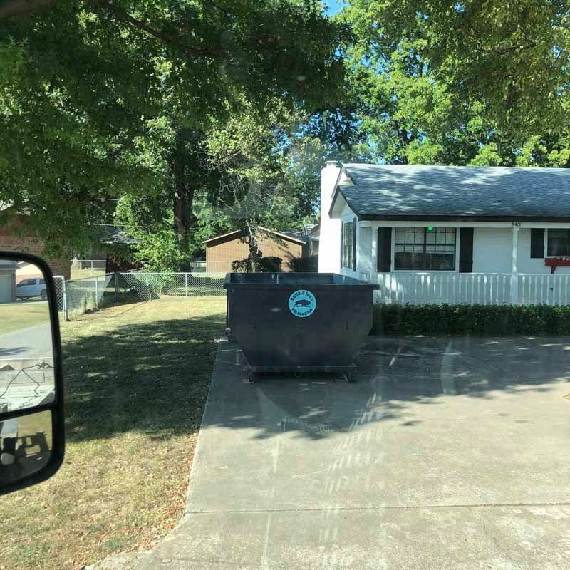 Black dumpster on a driveway beside a white house with trees and a fence in the yard