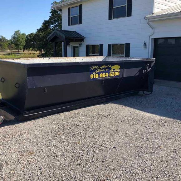 Large black dumpster on a gravel driveway beside a white house and garage, with yellow phone number lettering