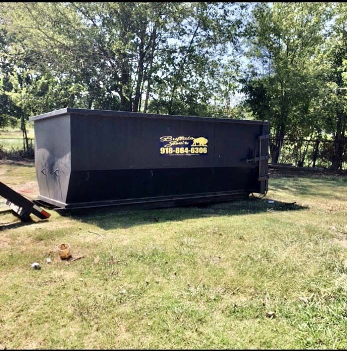 Large black dumpster on a grassy yard with trees in the background.