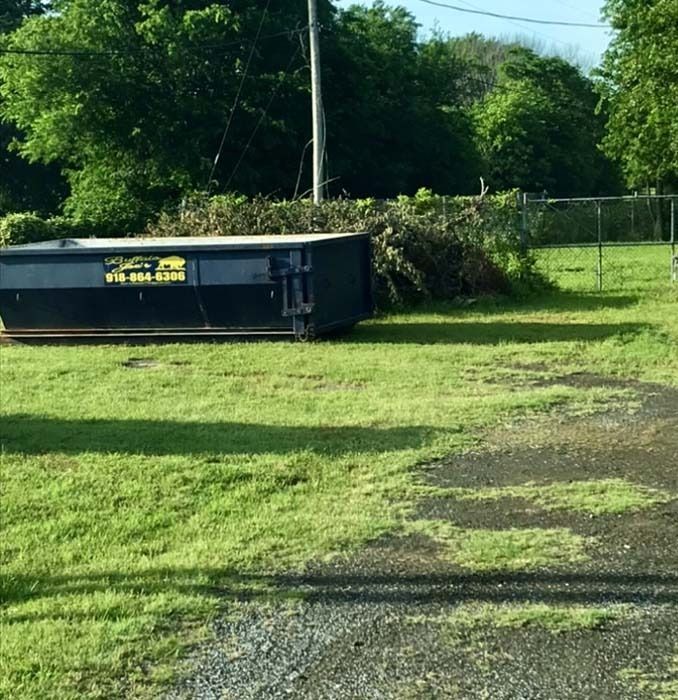 A black dumpster sits on a grassy field beside a chain-link fence and trees.