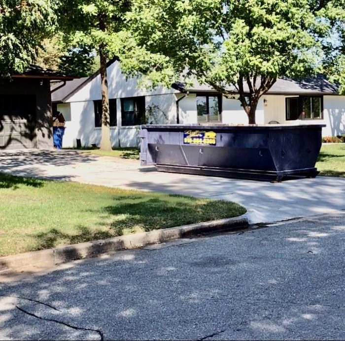 Large black dumpster on a driveway in front of a white building with trees and a grassy lawn.