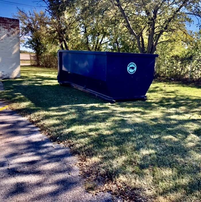 Blue dumpster on grass beside a building and trees in a sunny yard