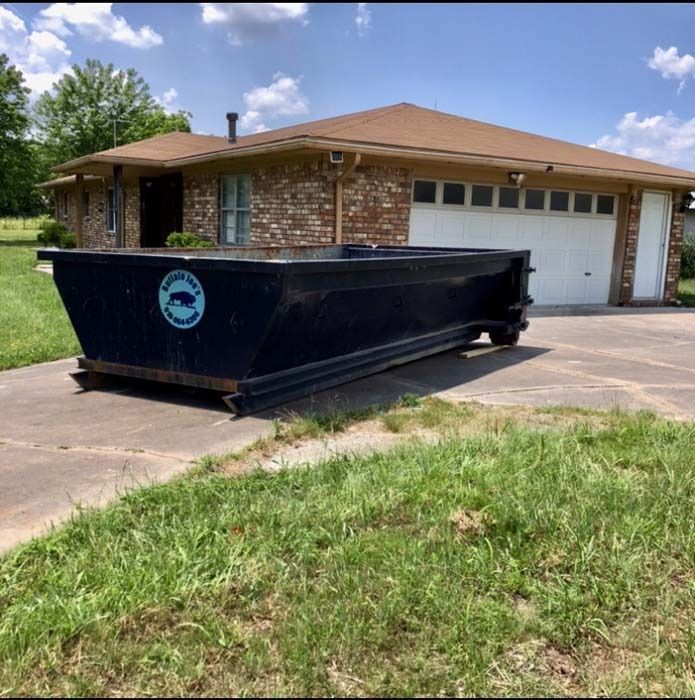 Large black dumpster in a driveway in front of a brick house on a sunny day