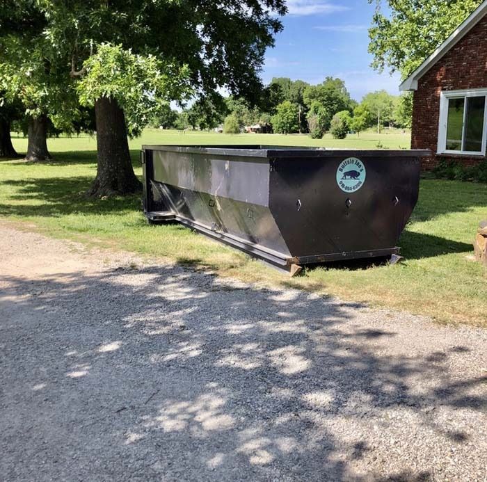 Large black dumpster on a gravel driveway beside a brick building and grassy yard with trees