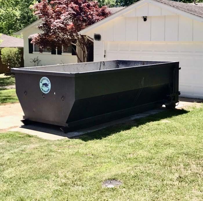 Black dumpster on a driveway in front of a white garage, with a grassy lawn and trees nearby