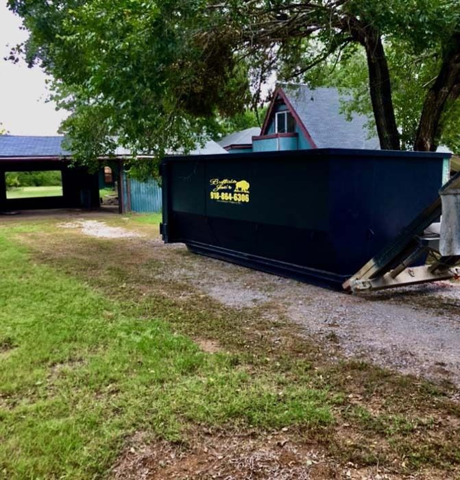 Black dumpster on gravel beside a house, with a grassy yard and trees around it.