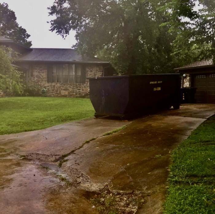 Dumpster blocking a wet driveway beside a stone house and garage in a grassy yard