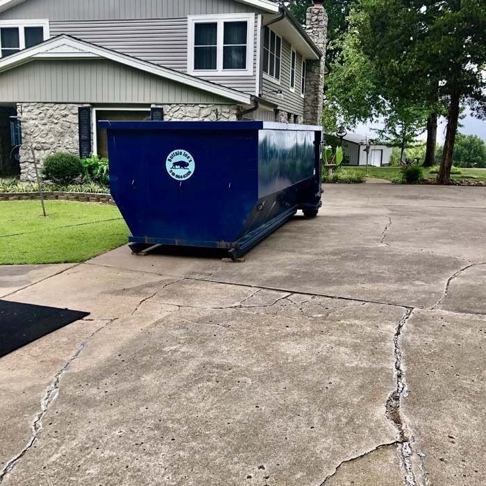 Blue dumpster on a cracked concrete driveway beside a house and lawn.