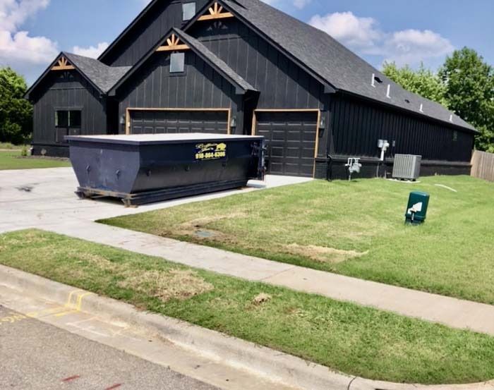 Black house with driveway, lawn, and large dumpster in front under a partly cloudy sky