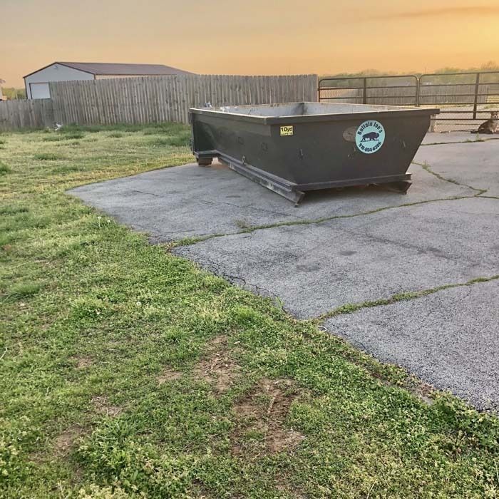 Large black dumpster on cracked pavement beside grass and a wooden fence at dusk
