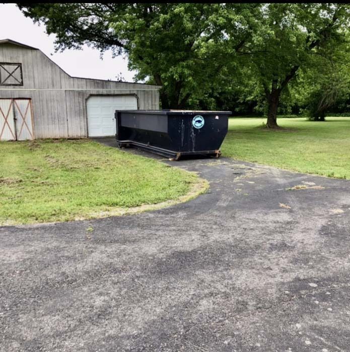 Black dumpster beside a white barn on a grassy lawn, next to a curved paved driveway under a large tree.