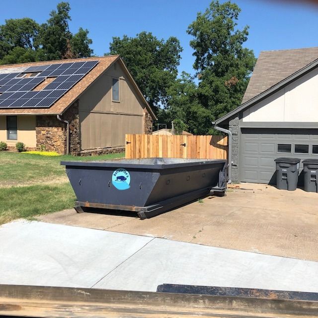 Large blue dumpster in a driveway beside a gray garage and fenced yard on a sunny day