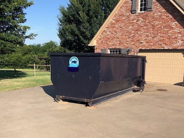 Large black dumpster on a driveway beside a brick garage and trees on a sunny day
