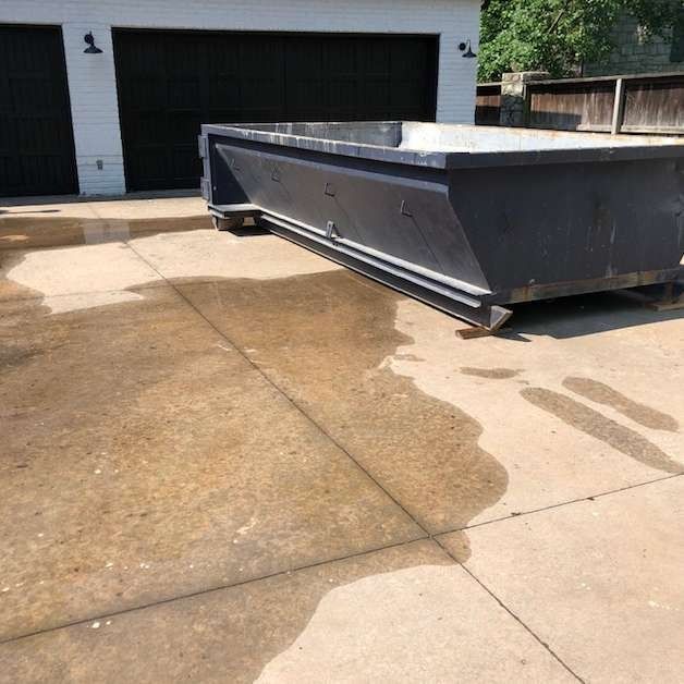 Large gray dumpster in a driveway beside a garage, with wet pavement and water stains.