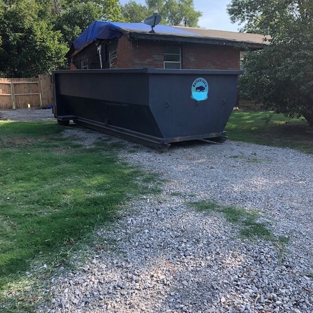 Black dumpster beside a brick building on a gravel driveway, with grass and trees around it