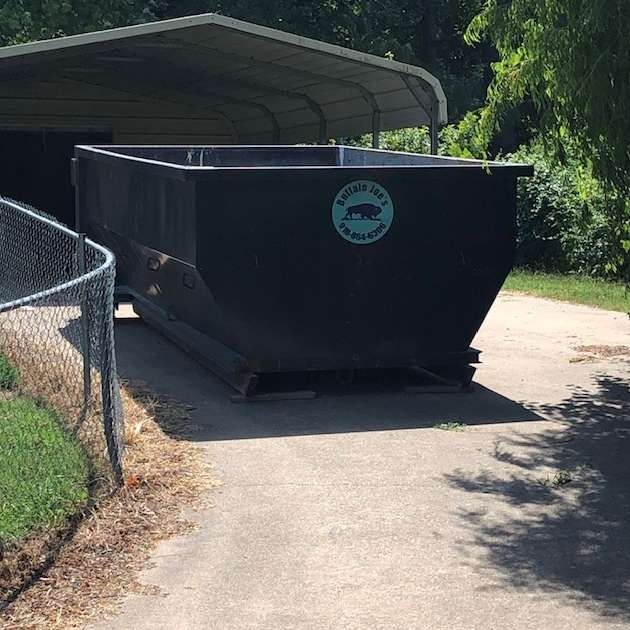 Large black dumpster on a paved path beside a chain-link fence and shaded trees
