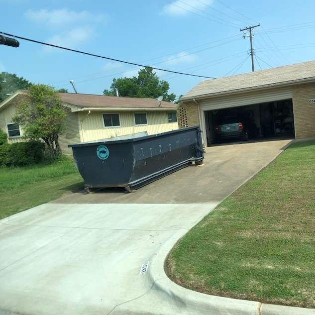 Large blue dumpster on a driveway beside a beige house with an open garage.