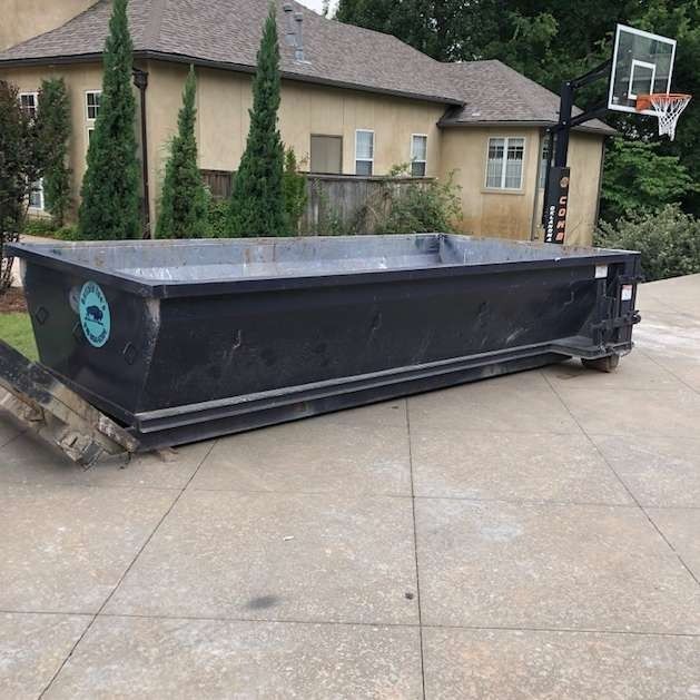 Large black dumpster bin on a driveway beside a house, with a basketball hoop in the background.