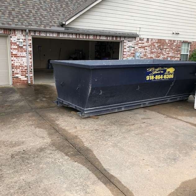 Large dark-blue dumpster parked on a driveway in front of a garage of a brick house.