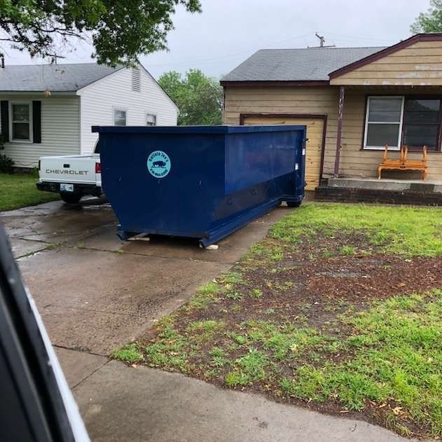Blue dumpster on a wet residential driveway beside a house and pickup truck.