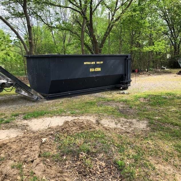 Black construction dumpster in a wooded yard beside a dirt path and grass
