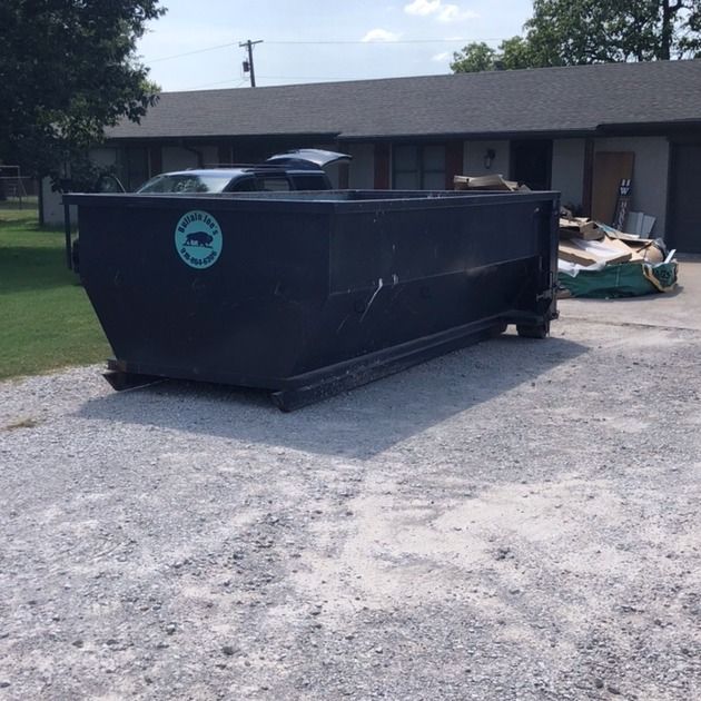 Large black dumpster on a gravel driveway beside a house, with construction debris piled nearby.