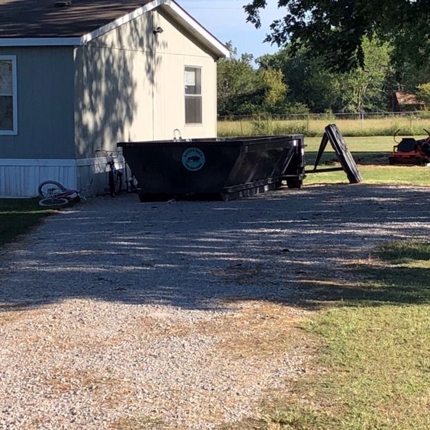 Gravel driveway beside a house with a black dumpster and a ladder in the yard