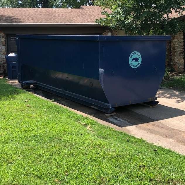 Large blue dumpster parked on a driveway beside a brick house and green lawn.