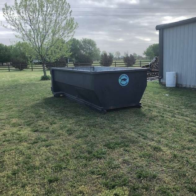 Black dumpster beside a gray metal building in a grassy yard under cloudy sky
