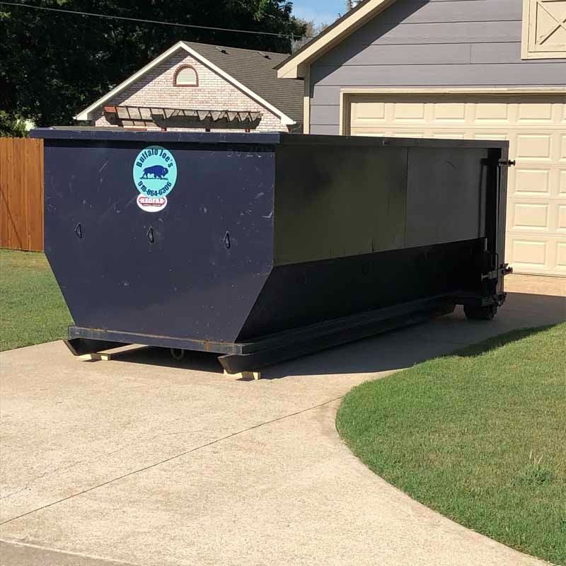 Large black dumpster on a driveway in front of a suburban house and garage.